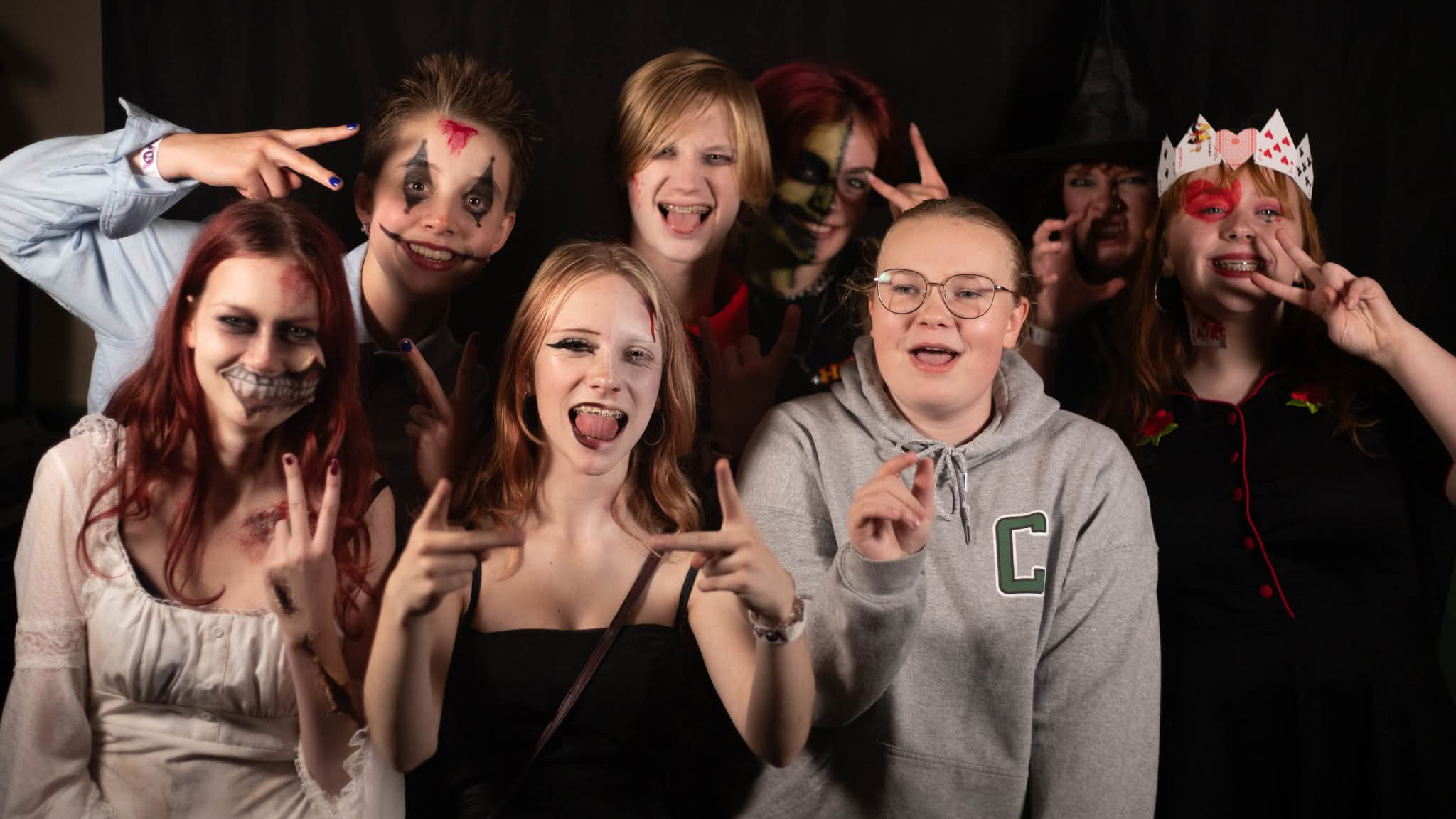 Groep jongeren met Halloween-schmink en kostuums poseert lachend voor de camera met handgebaren tegen zwarte achtergrond