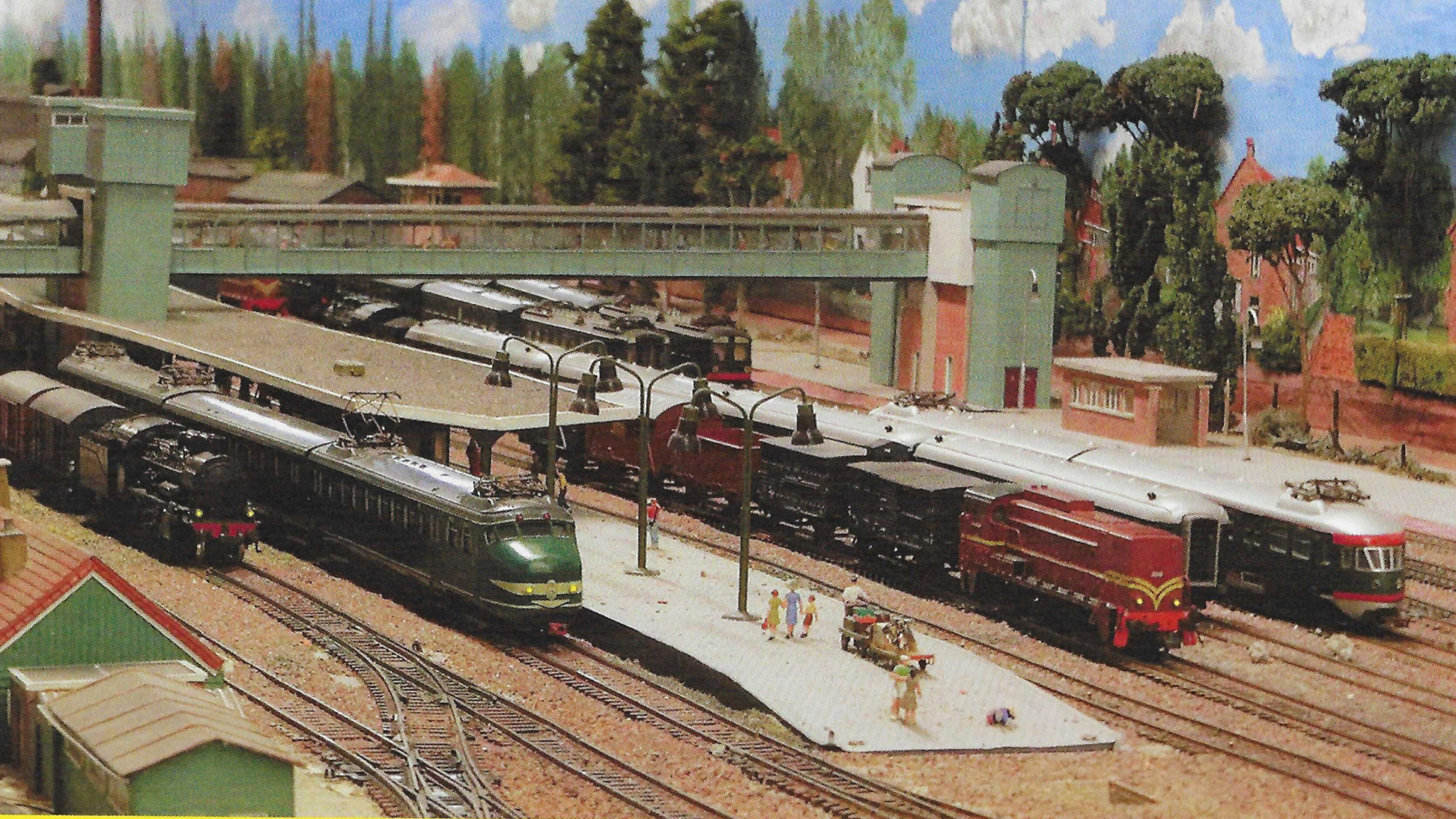 Modelspoor-treinstation met perrons, voetbrug en twee locomotieven, met kleine figuurtjes en bomen op de achtergrond