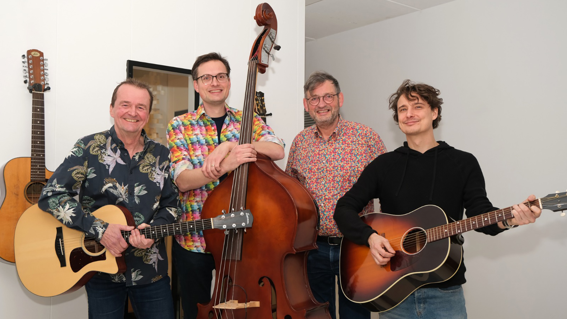 Vier muzikanten poseren in een studio met twee akoestische gitaren en een contrabas, met gitaren aan de muur