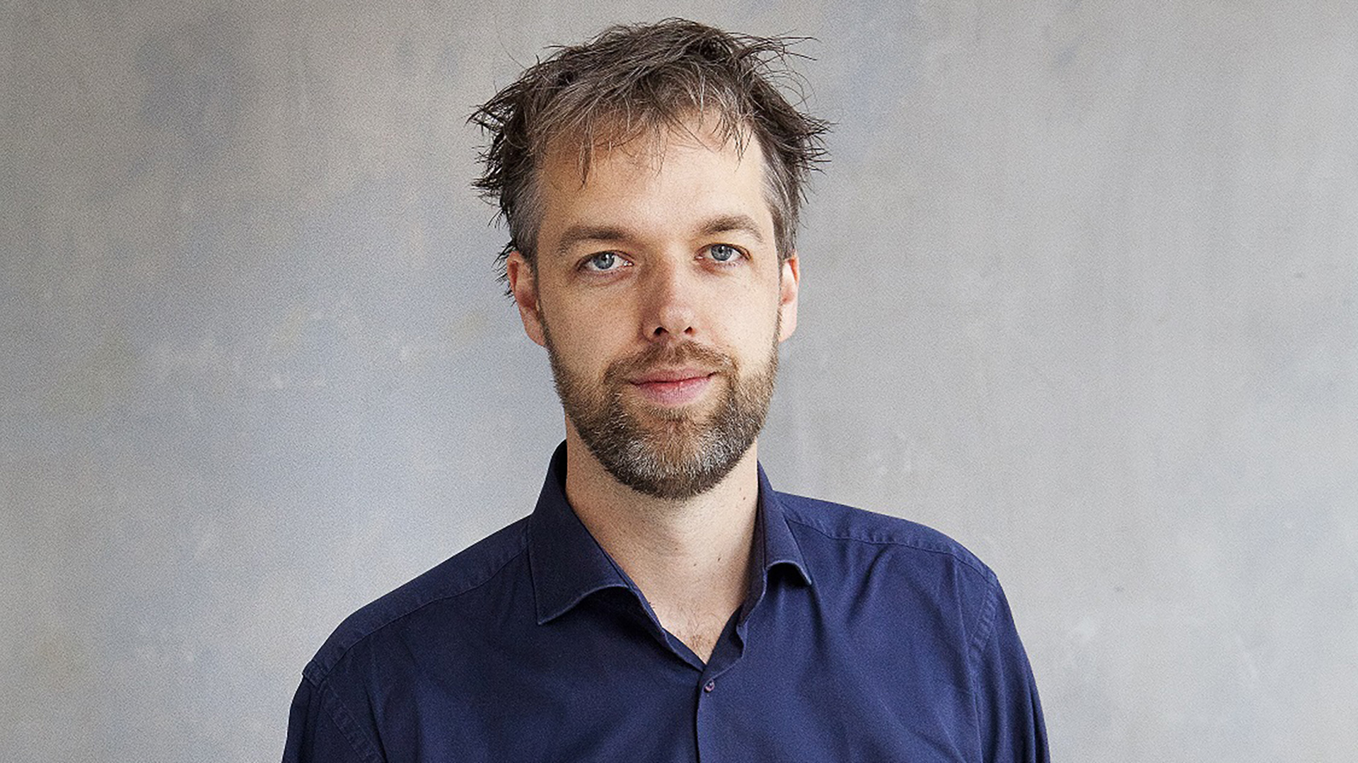 Portret van een man met kort rommelig haar en baard, in een donkerblauw overhemd tegen een grijze achtergrond