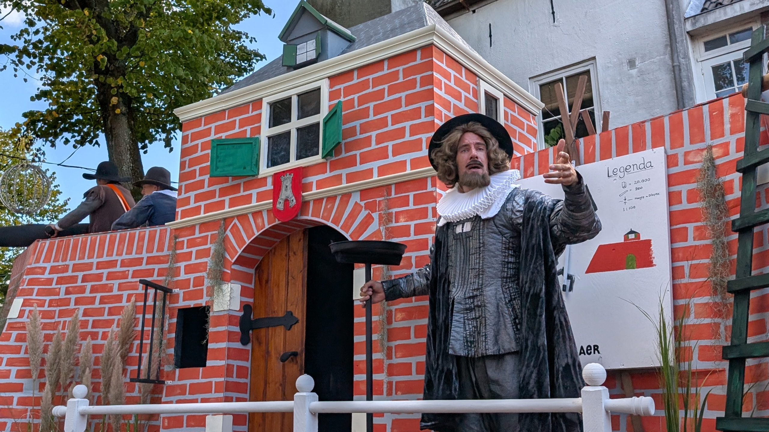 Man in historische kleding spreekt op een podium voor een decor van een bakstenen poortgebouw