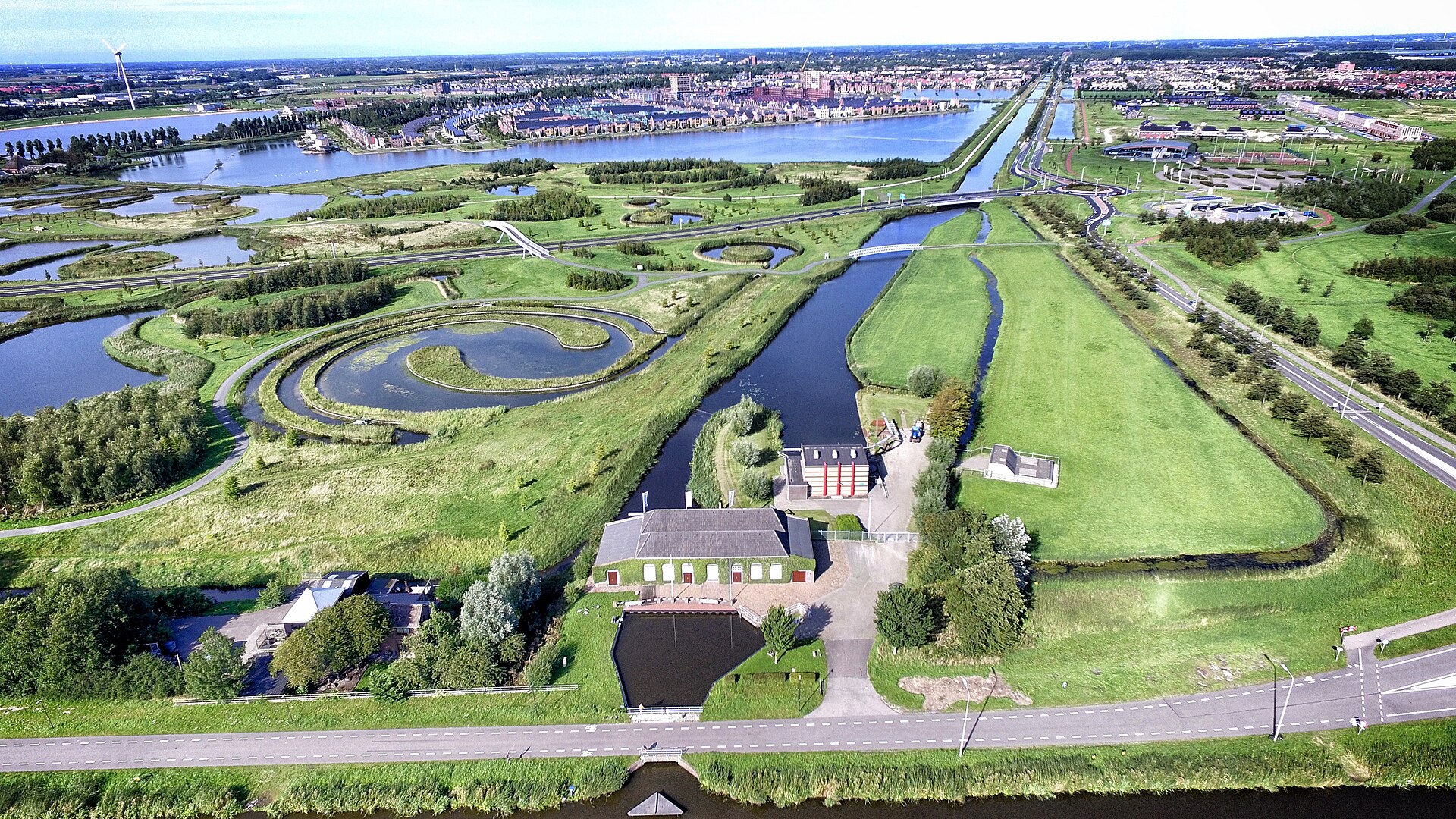 Luchtfoto van een waterlandschap met kanalen, wegen en een klein gemaal, met woonwijk en meren op de achtergrond
