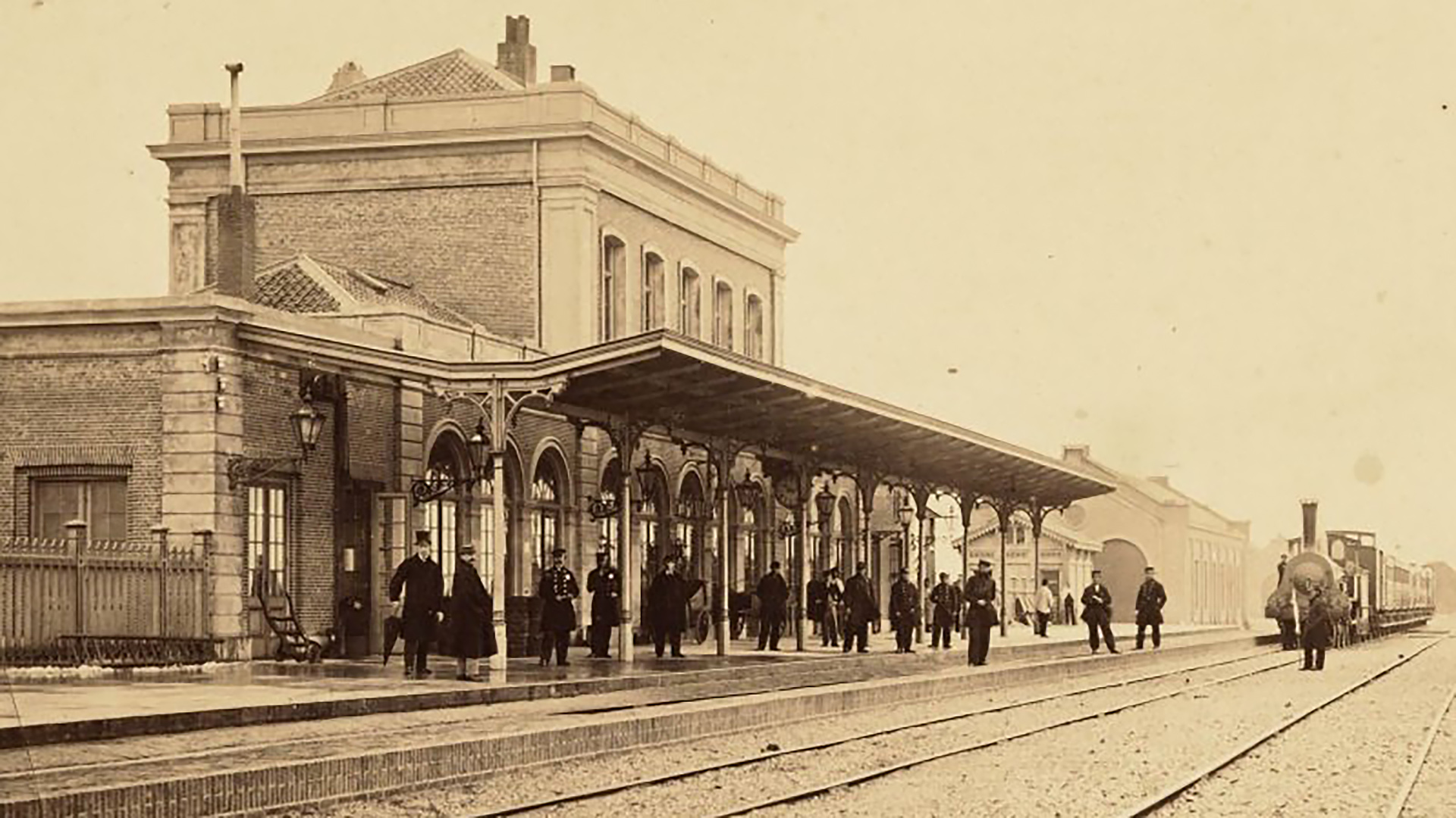 Historisch treinstation met overkapt perron, wachtende reizigers en een stoomlocomotief op de rails rechts