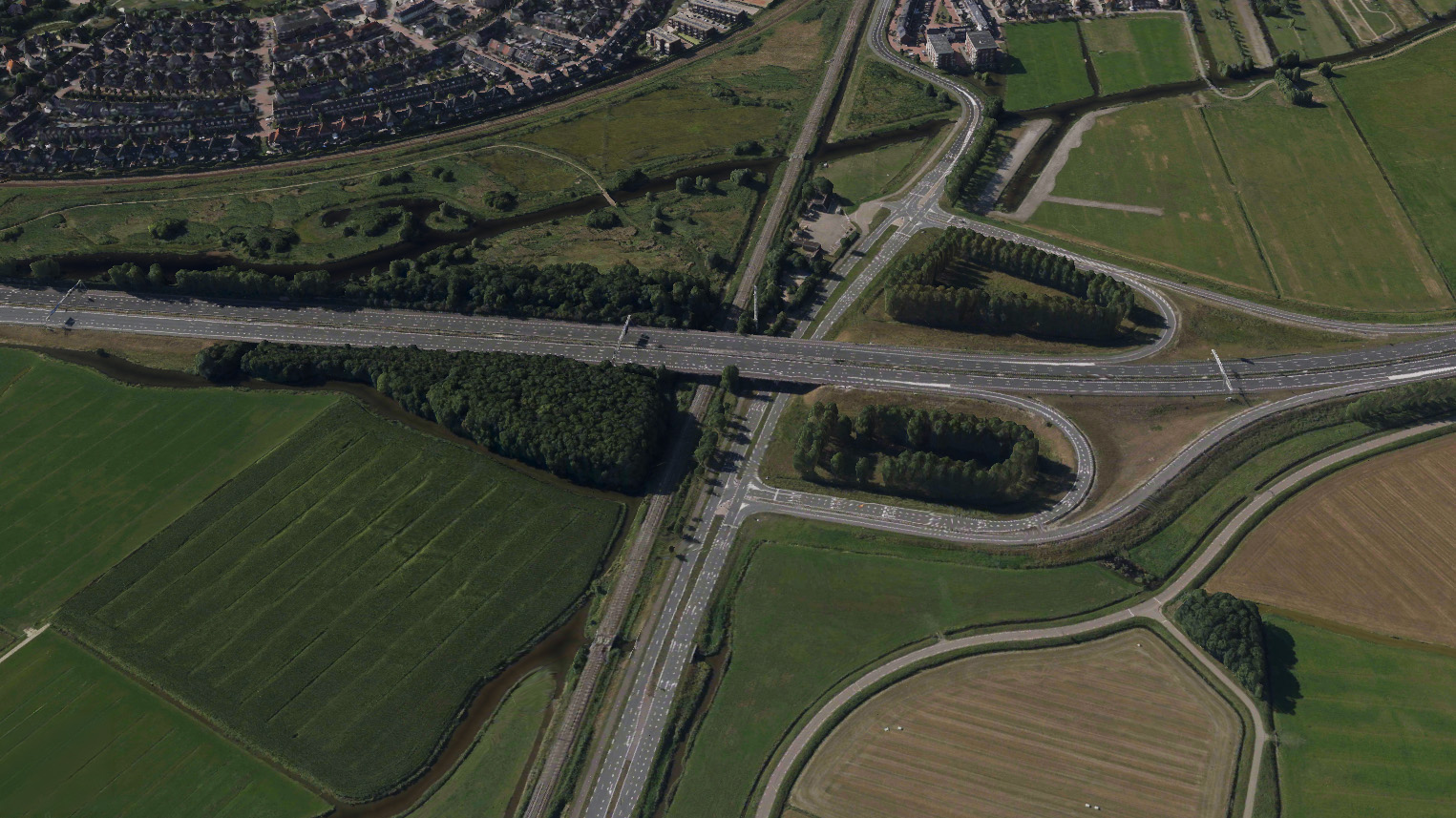 Luchtfoto van snelweg met verkeersknooppunt en viaduct over spoorlijn, omringd door velden, water en woonwijk