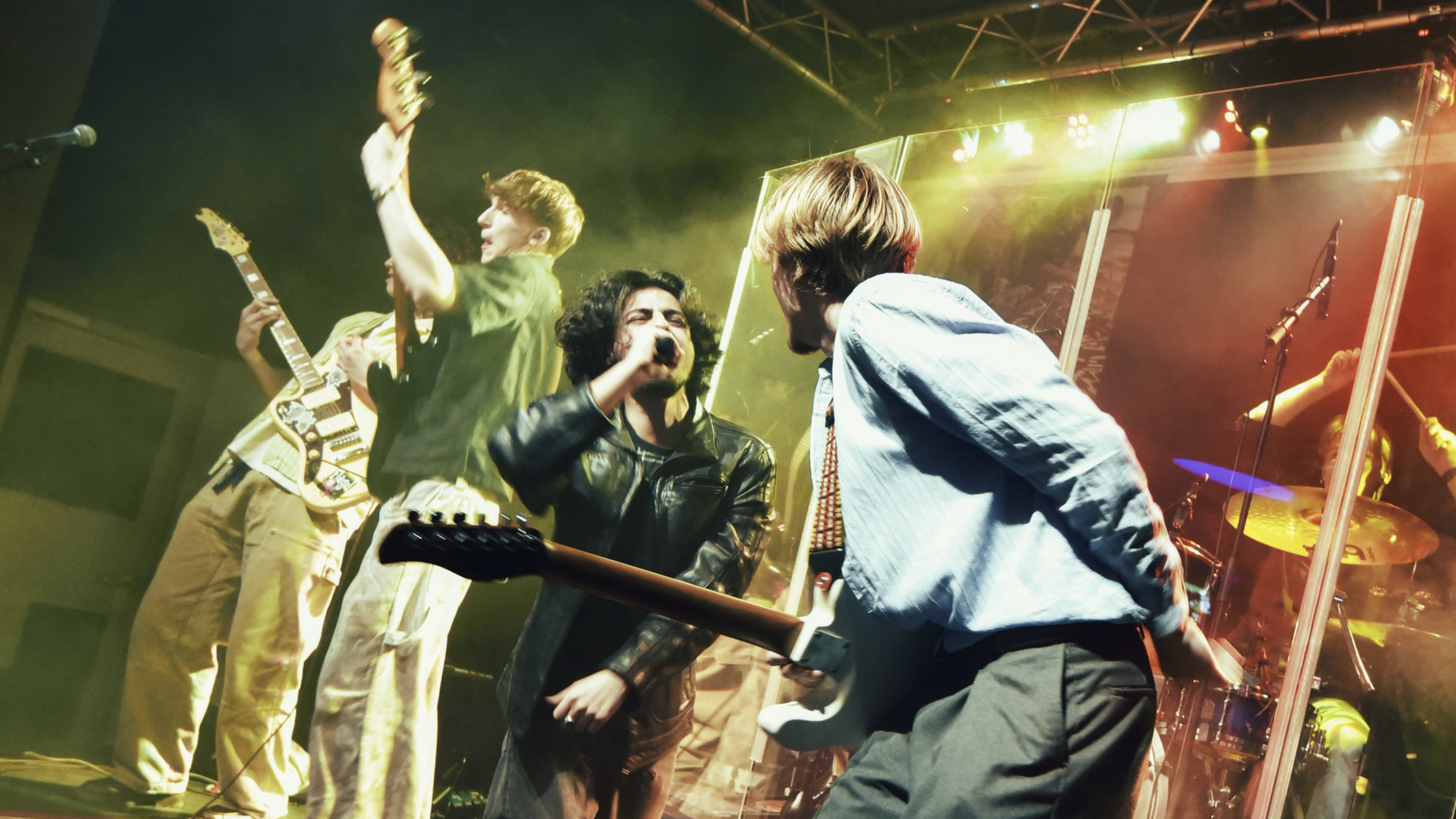 Rockband op het podium met zanger met microfoon en gitaristen, fel podiumlicht en drumstel achter plexiglas