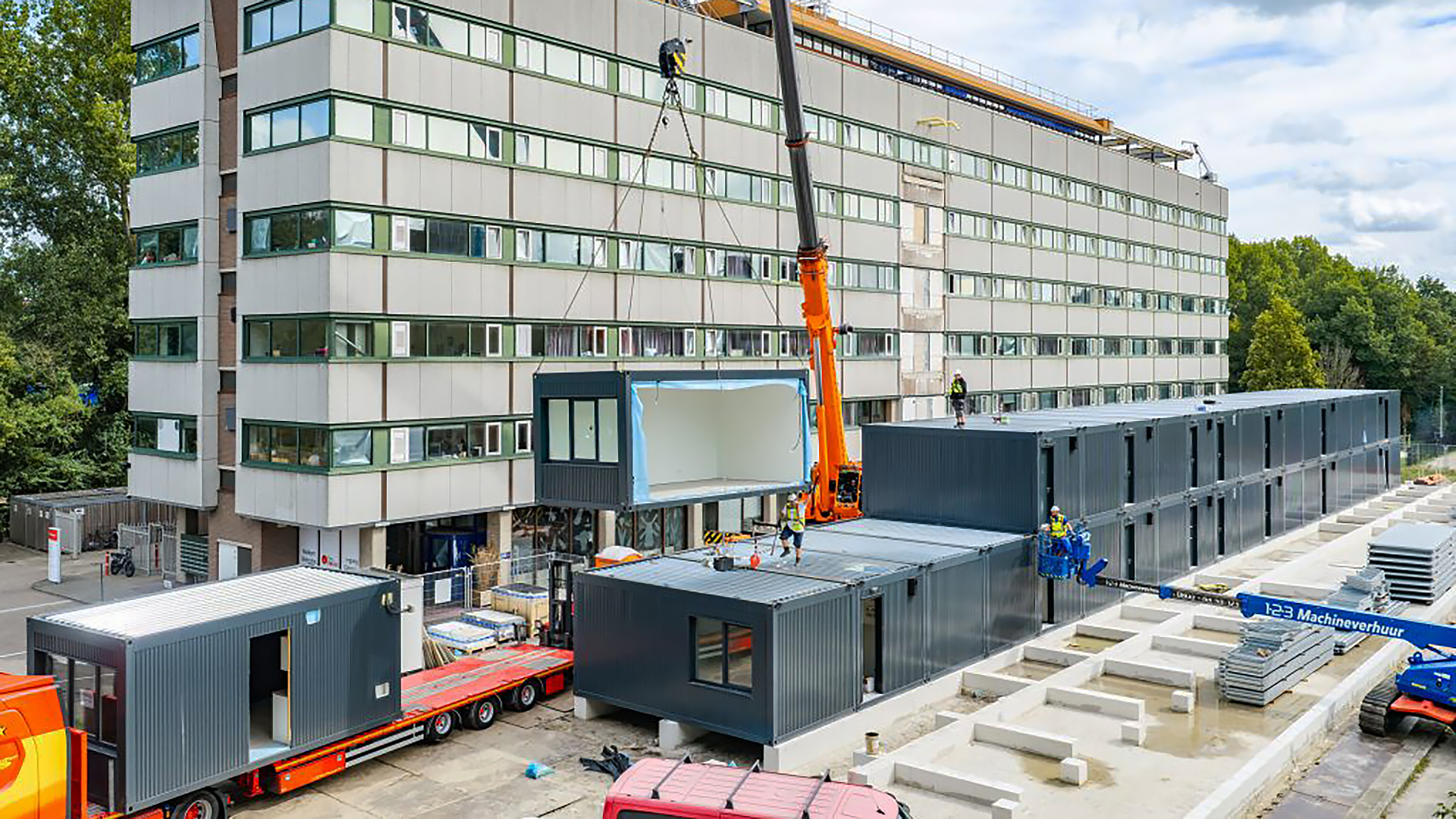 Bouwplaats met kraan die een modulaire unit plaatst naast een groot flatgebouw, met meerdere gestapelde containers en bouwvakkers