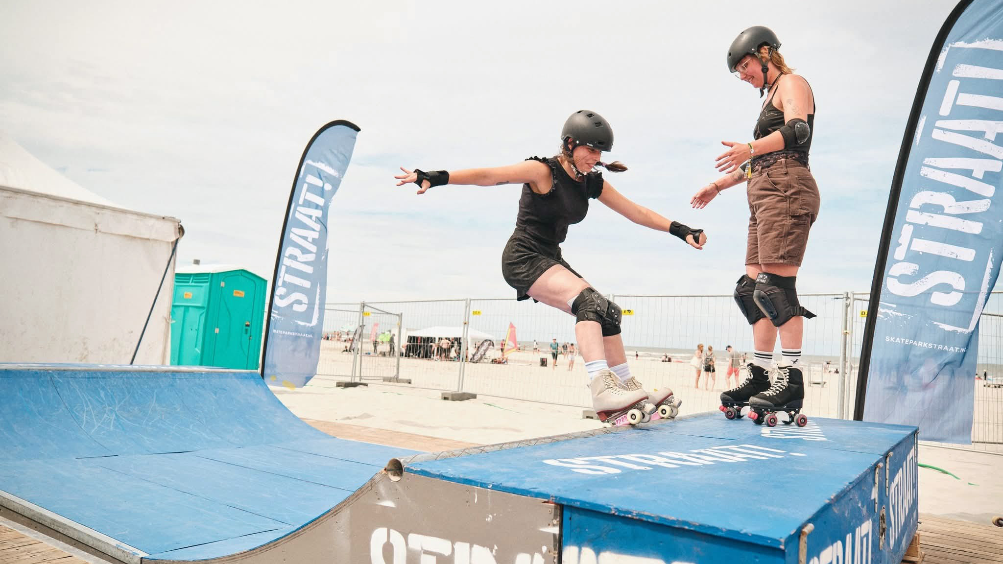 Twee rolschaatsers met helm op een blauwe ramp op het strand, met spandoeken met de tekst “STRAAT”