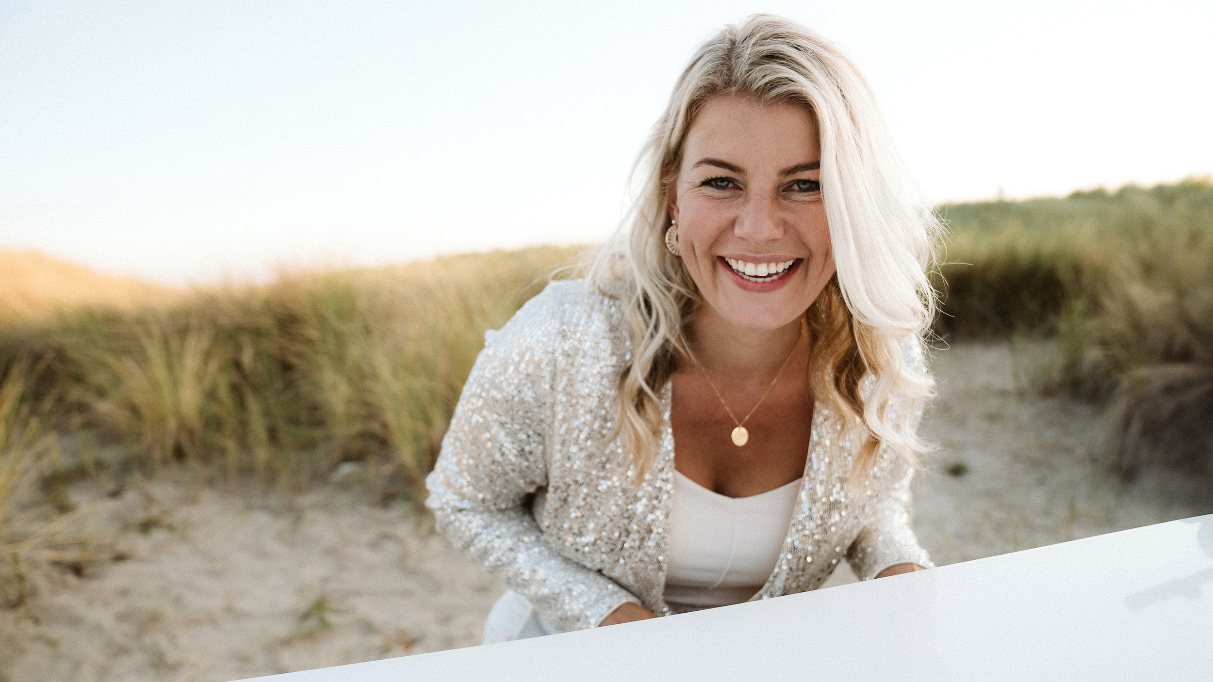 Lachende blonde vrouw in glinsterend jasje bij duinen en strand, kijkend in de camera bij zacht avondlicht